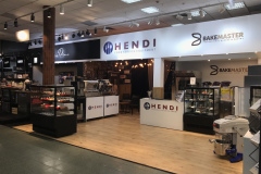 Nowoczesny salon wystawowy prezentuje sprzęt gastronomiczny Hendi i Bakemaster, z ladami, komercyjnymi urządzeniami kuchennymi i szklanymi gablotami wypełnionymi wypiekami w jasnym świetle. Szyldy z nazwami obu marek wiszą powyżej, a produkty są starannie ułożone na podłodze i półkach. projekt i nadzór Tatjana Kobuszewska trksystem.com