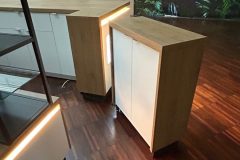 Drewniano-biała szafka z podświetlanym panelem stoi na wypolerowanej drewnianej podłodze w nowoczesnym wnętrzu Manufaktury Łódź, obok lady i szklanej witryny, z liściastą tapetą i sprzętem przeciwpożarowym w tle. projekt i nadzór Tatjana Kobuszewska trksystem.com