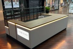 Nowoczesna wyspa handlowa "Słoik Home" o powierzchni z jasnego drewna i podświetlanych krawędziach stoi w łódzkiej Manufakturze. Na biurku stoi roślina doniczkowa, przywołująca na myśl domowy klimat. Projekt i nadzór Tatjana Kobuszewska trksystem.com