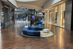 Nowoczesne centrum handlowe z drewnianymi podłogami, zakrzywioną architekturą, szklanymi balustradami i kilkoma zamkniętymi sklepami. W centrum znajdują się niebieskie siedzenia i kolorowe dywany, a w tle widoczne są ruchome schody i świetliki. projekt i nadzór Tatjana Kobuszewska trksystem.com