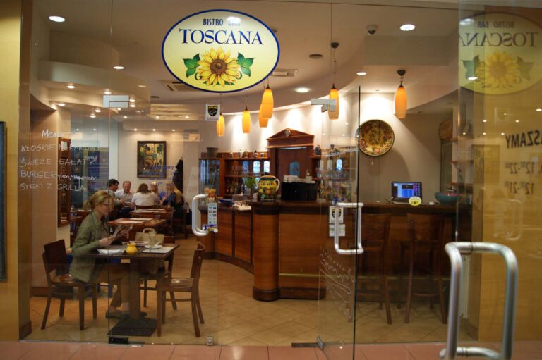 Szklane drzwi prowadzą do Toscana Bistro Bar, przytulnego miejsca z ciepłym oświetleniem, drewnianymi stołami i zakrzywionym barem. Kilka osób siedzi i je. Menu na ścianie zawiera listę włoskich dań w języku polskim. Z sufitu zwisają żółte lampy wiszące. projekt i nadzór Tatjana Kobuszewska trksystem.com