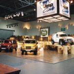 Kryty pokaz samochodowy w Stoisku Jeep na Expo XXI obejmuje cztery pojazdy Jeep w różnych kolorach na drewnianej platformie. Powyżej wisi duży napis "Jeep Club Polska" z napisem ONLY IN A JEEP, drewnianym wystrojem i jasnym oświetleniem w tle. projekt i nadzór Tatjana Kobuszewska trksystem.com