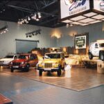 W Stoisku Jeep na Expo XXI cztery pojazdy Jeep są prezentowane na wytrzymałych, podwyższonych platformach otoczonych skałami i drewnianym ogrodzeniem. Reflektory podświetlają Jeepy, a nad nimi znajdują się banery i znaki firmowe. Przy lewej krawędzi stoi człowiek. projekt i nadzór Tatjana Kobuszewska trksystem.com