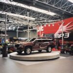 Bordowy SUV z brandingiem JVC stoi na okrągłej platformie w muzycznym stoisku JVC w hali wystawowej. Za nim, duży czerwony baner JVC podkreśla "MP3". projekt i nadzór Tatjana Kobuszewska trksystem.com