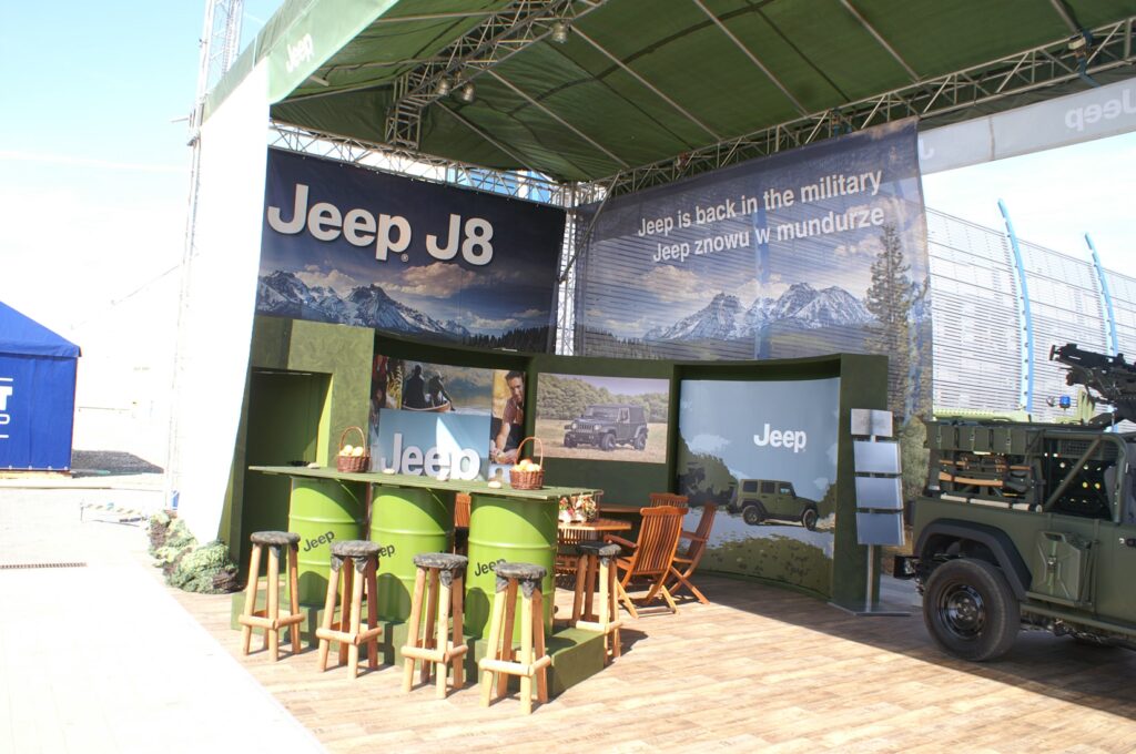 Stoisko Jeep na targach MSPO Kielce - zaczepno-odporne; zewnętrzne stoisko wystawiennicze pod zadaszeniem dla Jeepa J8, z banerami z górami i pojazdami wojskowymi, zielonymi ladami, stołkami barowymi, drewnianymi krzesłami i częścią militarnej gamy Jeepa po prawej stronie. projekt i nadzór Tatjana Kobuszewska