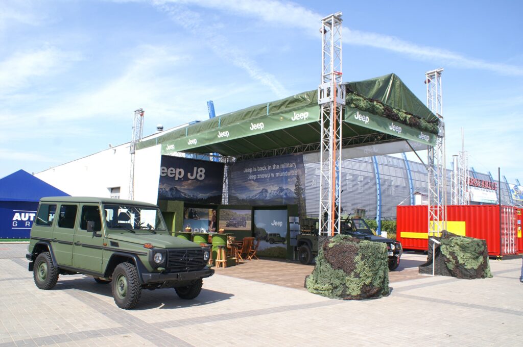 Zielony Jeep J8 i zakamuflowany pojazd są zaparkowane na zewnątrz stoiska Jeep na targach MSPO Kielce - zaczepno-odporne, stoisko o tematyce wojskowej z zadaszeniem, banerami i krzesłami. Jasne niebo, pobliskie kontenery i budynki otaczają wystawę. projekt i nadzór Tatjana Kobuszewska