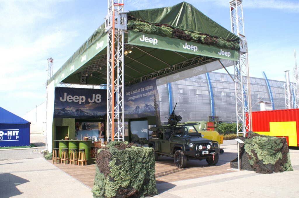 Zielony pojazd wojskowy Jeep J8 z zamontowaną na dachu bronią jest prezentowany na stoisku Jeep na targach MSPO. Na stoisku znajduje się zielona siatka maskująca, banery, stołki barowe, stoły i górskie tła na zewnątrz na utwardzonym terenie. projekt i nadzór Tatjana Kobuszewska
