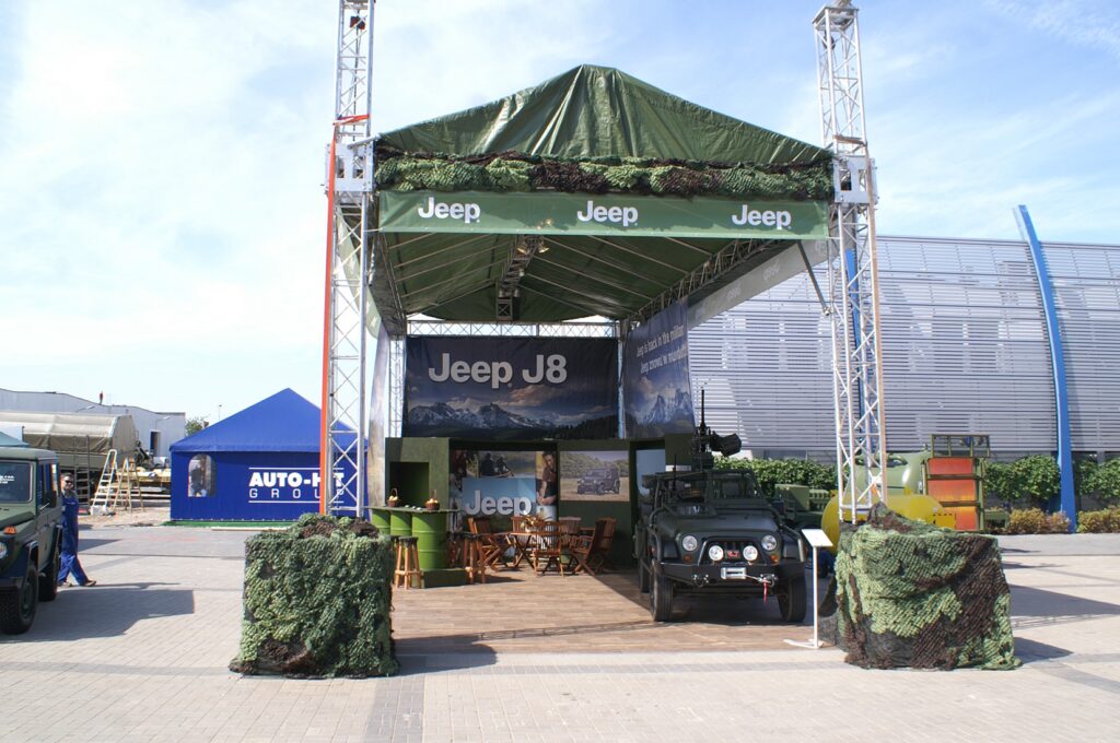 Zewnętrzna wystawa Jeepa pod zadaszeniem z logo "Jeep", siatką kamuflażową i pojazdem Jeep w stylu wojskowym z przodu. Stoisko Jeep na targach MSPO Kielce - zaczepno-odporne, prezentuje militarną gamę jeepów na utwardzonej działce z niebieskim namiotem w tle. projekt i nadzór Tatjana Kobuszewska