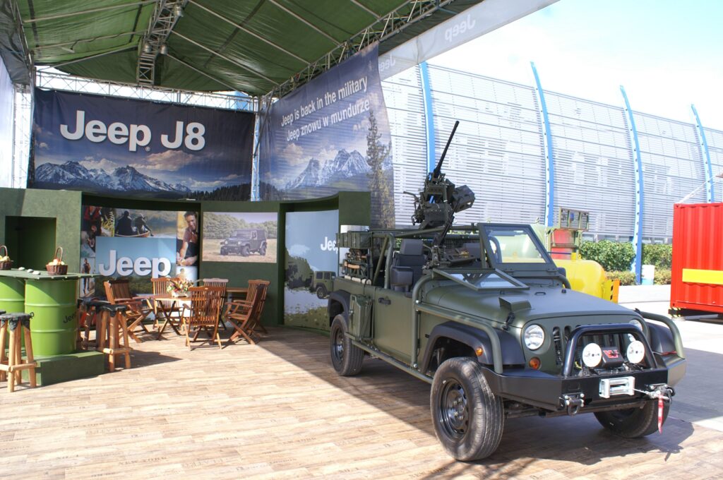Ciemnozielony wojskowy Jeep J8, reprezentujący militarną gamę Jeep, z zamontowanym sprzętem stoi na drewnianej podłodze pod otwartym namiotem. Stoisko Jeep na targach MSPO Kielce zdobią outdoorowe banery i wygodna strefa siedzenia. projekt i nadzór Tatjana Kobuszewska