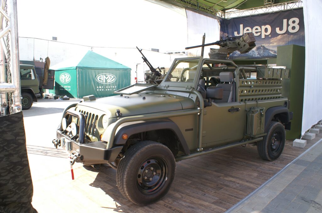 Wojskowy Jeep J8 z oliwkowo-zielonym lakierem jest wystawiony w Jeep stoisko na targach MSPO. Nie ma drzwi ani dachu, ma zamontowane uzbrojenie na górze, wzmocnione zderzaki, wytrzymałe opony i bagażniki. W tle znajdują się namioty i banery z logo. projekt i nadzór Tatjana Kobuszewska