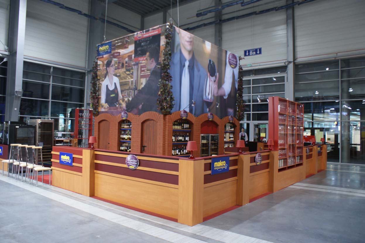 Przestronne, dobrze oświetlone stoisko targowe Makro Polagra z drewnianymi ladami i półkami eksponującymi butelki. Duże banery powyżej przedstawiają ludzi wchodzących w interakcje w sklepie. Stoisko jest udekorowane girlandami i markowymi znakami "makro" w hali wystawienniczej. projekt i nadzór Tatjana Kobuszewska trksystem.com