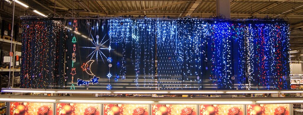 Na ciemnym tle hali Makro z sufitami w stylu industrialnym rozstawiono wiszące świąteczne lampki w różnych kolorach i kształtach, w tym gwiazdki, księżyce i płatki śniegu. Ciepłe światło rozchodzi się z podświetlanych paneli wzdłuż dolnej krawędzi. projekt i nadzór Tatjana Kobuszewska