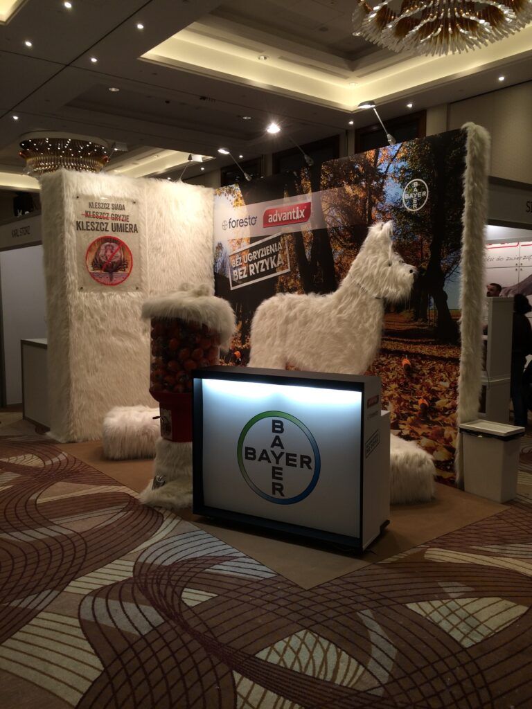 Stoisko targowe ozdobione sztucznym białym futrem, z dużą figurką lamy i automatem do gumy do żucia. Logo Bayer znajduje się na ladzie frontowej, z banerami o tematyce jesiennej — projekt i nadzór Tatjana Kobuszewska trksystem.com. projekt i nadzór Tatjana Kobuszewska trksystem.com