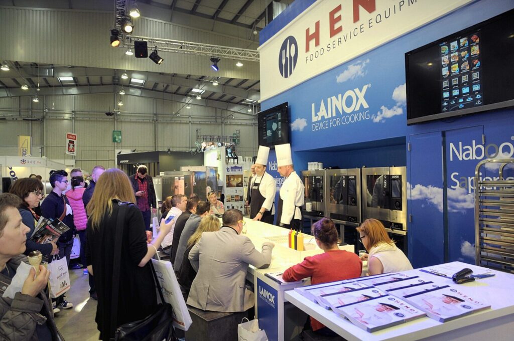 Tłum ogląda dwóch kucharzy w białych kapeluszach, którzy prezentują pokaz gotowania na stoisku targowym sprzętu gastronomicznego Lainox i Henny. W otoczeniu znajdują się urządzenia kuchenne i ekspozycje promocyjne autorstwa projekt i nadzór Tatjana Kobuszewska trksystem.com. projekt i nadzór Tatjana Kobuszewska trksystem.com