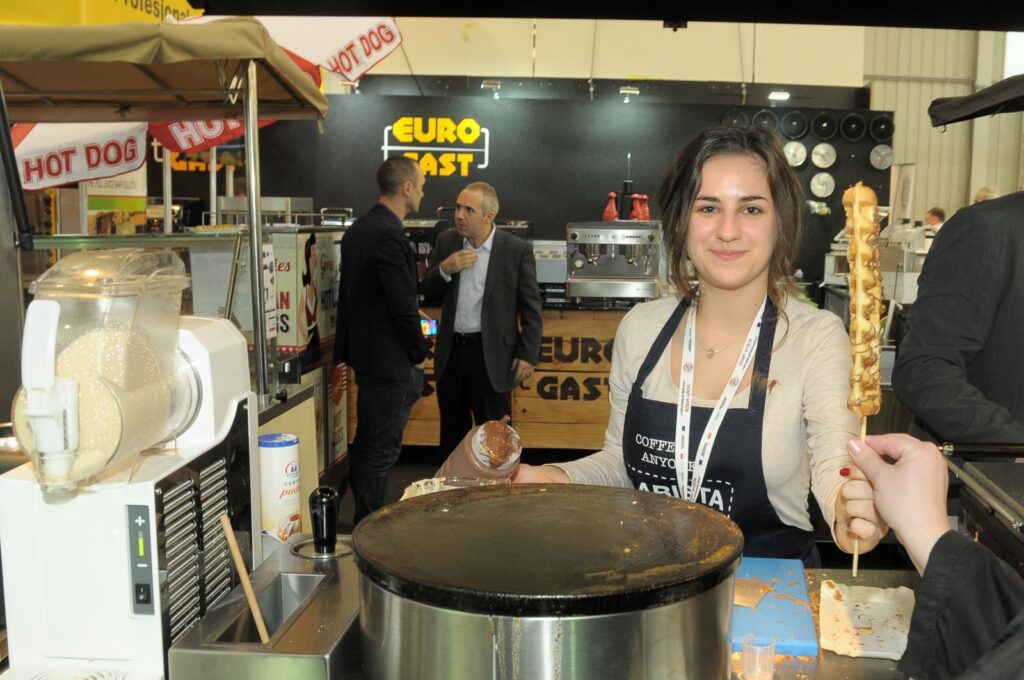 Podczas imprezy Eurogastro 2015 na stoisku gastronomicznym Eurogast kobieta w fartuchu trzyma na szpikulcu przekąskę polaną sosem. Uśmiecha się do klienta, a za nią widać sprzęt do przygotowywania naleśników oraz szyld „Euro Gast”. W tle rozmawiają dwaj mężczyźni, a nad nimi wisi szyld z hot dogiem, oddający żywą atmosferę stoiska „na szybko” –. projekt i nadzór Tatjana Kobuszewska