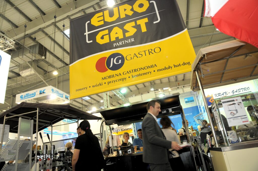 Ludzie spacerują i stoją w pobliżu dużego napisu „EURO GAST” oraz stoisk gastronomicznych na targach w hali, co przypomina tętniącą życiem atmosferę z wydarzenia 2015-eurogastro-eurogast-stoisko-na-szybko-27. W tym miejscu panuje ożywiony ruch – pod jasnym oświetleniem wiszą różne stoiska, sprzęt i banery reklamujące usługi cateringowe i gastronomiczne. projekt i nadzór Tatjana Kobuszewska