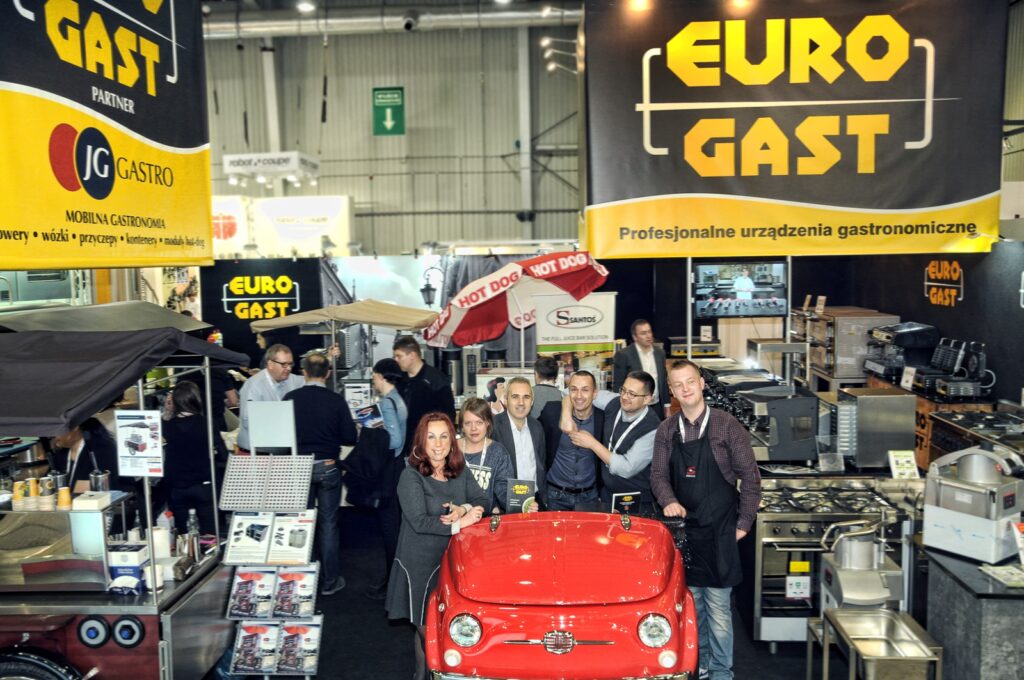 Grupa uśmiechniętych dorosłych stoi za małym czerwonym samochodem przy ruchliwym stoisku targowym „2015-eurogastro-stoisko-targowe-na-szybko” na targach Euro Gast, na którym prezentowany jest sprzęt kuchenny oraz duże banery w języku polskim. Pozostałą część jasno oświetlonej przestrzeni wystawowej wypełniają inni ludzie oraz urządzenia. projekt i nadzór Tatjana Kobuszewska