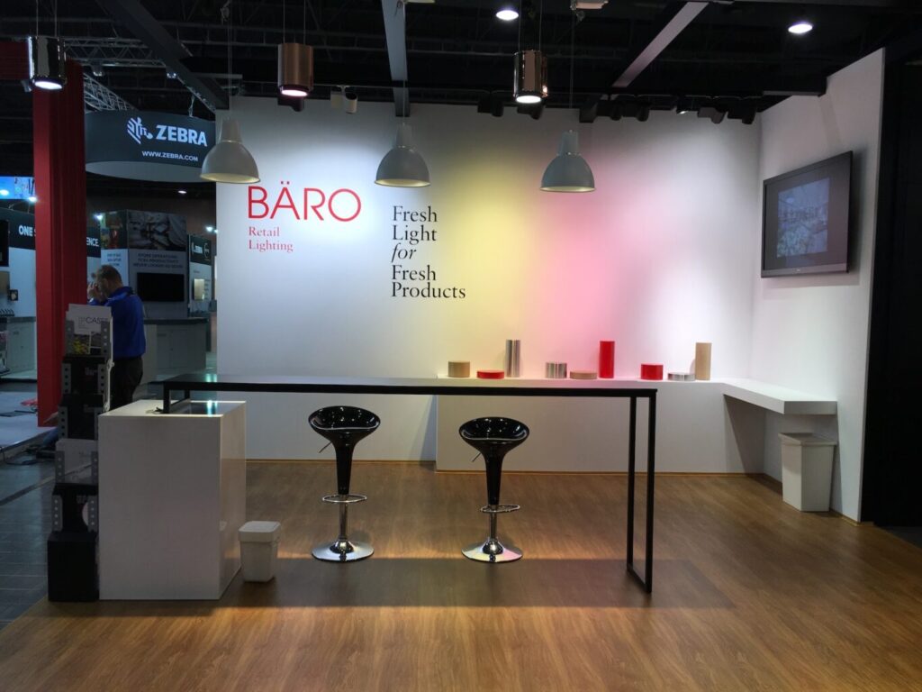Nowoczesne stoisko wystawowe firmy BÄRO Retail Lighting na targach Baro Retail Show 016, z białą ścianą z hasłem „Świeże światło dla świeżych produktów”, dwoma czarnymi hokerami barowymi przy długim barze, wiszącymi lampami nad ladą oraz kolorowymi cylindrami na wystawie. projekt i nadzór Tatjana Kobuszewska trksystem.com