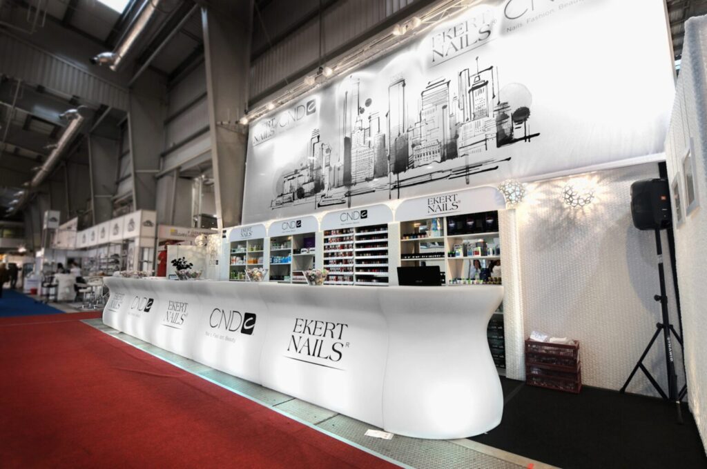 Nowoczesne stoisko salonu paznokci z zakrzywionym białym blatem, na którym widnieją logo „EKERT NAILS” i „CND”, półki z produktami do paznokci i ilustracja panoramy miasta. Projekt i nadzór Tatjana Kobuszewska trksystem.com. Znajduje się w dużej hali wystawowej. projekt i nadzór Tatjana Kobuszewska trksystem.com