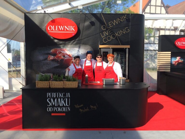 Cztery uśmiechnięte osoby w czerwonych fartuchach stoją za ladą ze świeżymi ziołami na wydarzeniu Olewnik live cooking, prezentującym projekt i nadzór Tatjana Kobuszewska trksystem.com. W tle czarno-czerwone oznakowanie i sprzęt kuchenny. projekt i nadzór Tatjana Kobuszewska trksystem.com