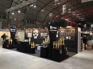 Stoisko targowe projektu i nadzoru Tatjany Kobuszewskiej (trksystem.com) eksponuje liczne złote i srebrne trofea, medale i nagrody na czarnych stojakach. Loga firm i certyfikaty zdobią ściany w dużej hali centrum kongresowego. projekt i nadzór Tatjana Kobuszewska trksystem.com