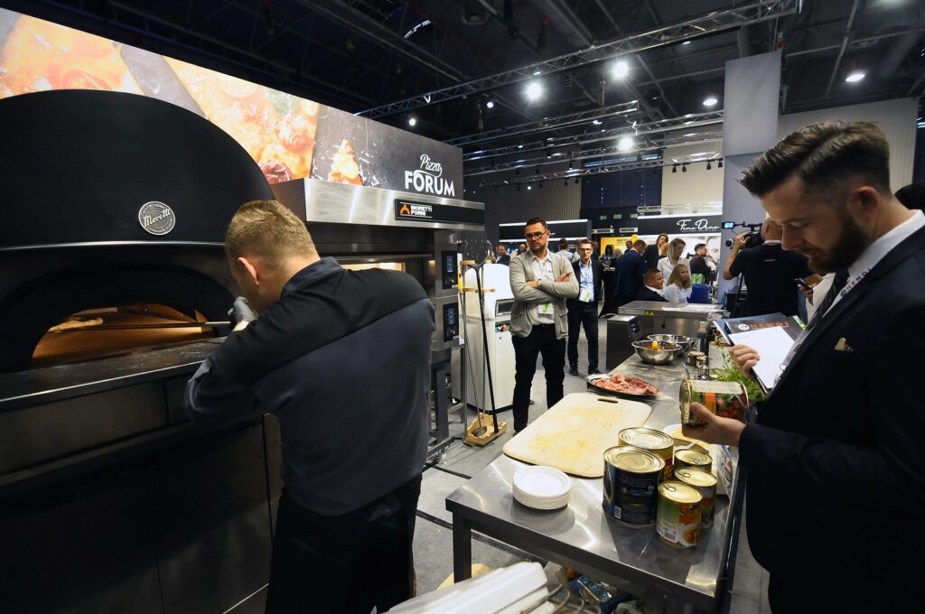 Podczas kongresu szefów kuchni i restauratorów Hendi 2017 mężczyzna w ciemnym stroju przygotowuje pizzę w dużym piecu, podczas gdy inny, ubrany w garnitur, sprawdza składniki w puszkach. Goście nawiązują kontakty towarzyskie i obserwują wydarzenia w nowoczesnej, dobrze oświetlonej sali. projekt i nadzór Tatjana Kobuszewska