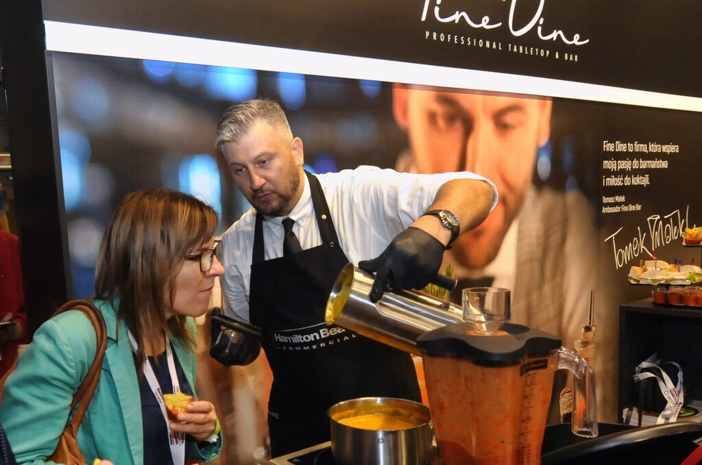 Kucharz w ciemnym fartuchu i rękawiczkach nalewa jasnopomarańczowy koktajl z blendera do kubka dla kobiety w okularach przy stoisku „Fine Vine”, na którym widnieją napisy i starannie ułożone kubki z próbkami. projekt i nadzór Tatjana Kobuszewska