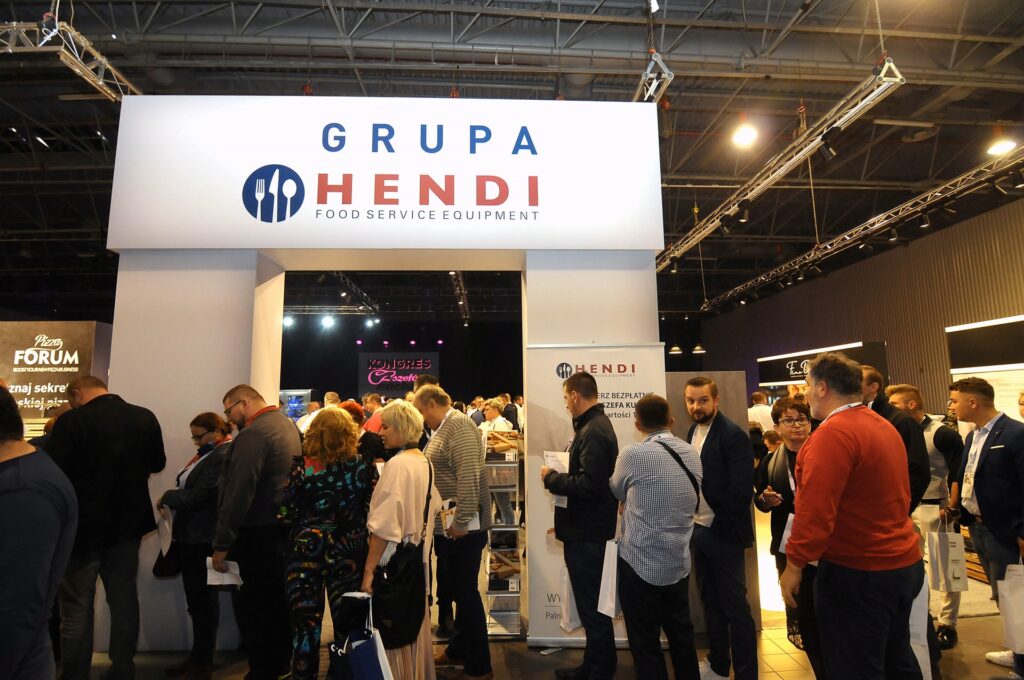 Tłum ludzi stoi i przechodzi obok bramy wejściowej do stoiska firmy Hendi na kongresie szefów kuchni i restauratorów, przy której widnieje napis „Grupa Hendi Food Service Equipment”, w jasno oświetlonej hali wystawowej wypełnionej banerami i ekspozycjami. projekt i nadzór Tatjana Kobuszewska