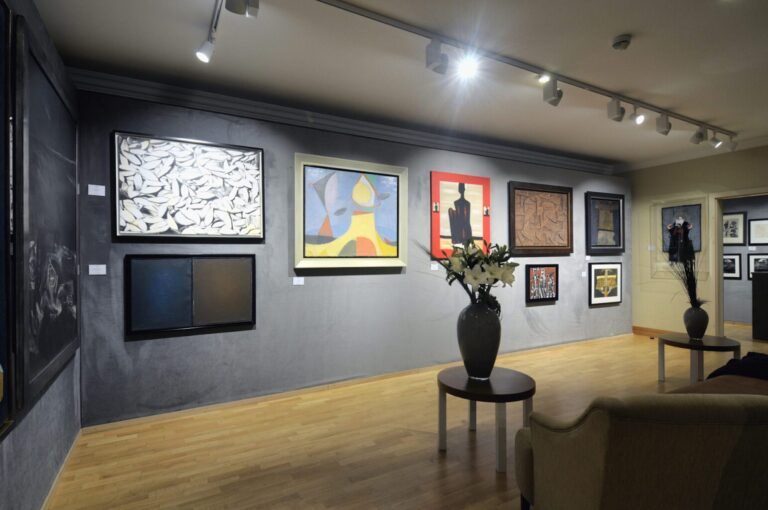 Nowoczesna galeria sztuki z obrazami na szarych ścianach, drewnianą podłogą, okrągłym stołem z wazonem z białymi kwiatami i beżową sofą pod reflektorami — projekt i nadzór Tatjana Kobuszewska trksystem.com. projekt i nadzór Tatjana Kobuszewska trksystem.com