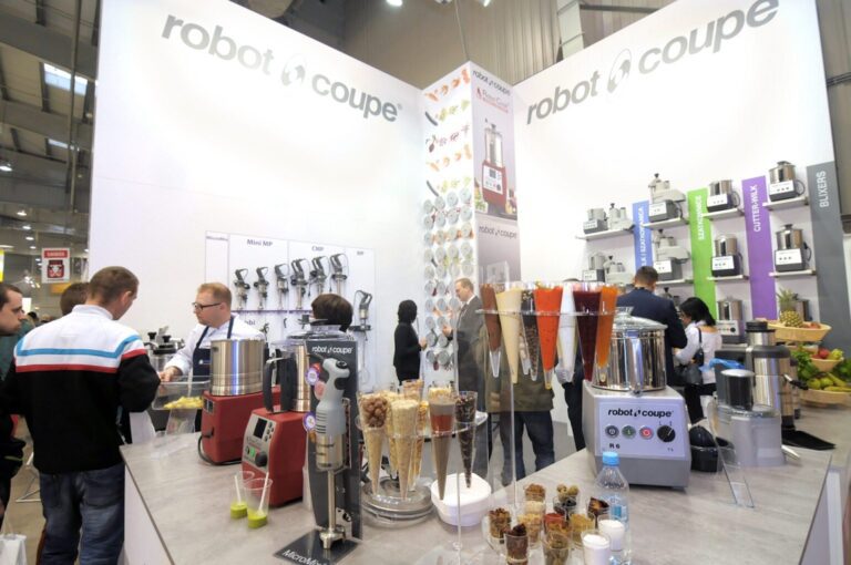 Stoisko targowe prezentuje sprzęt kuchenny Robot Coupe. Odwiedzający wchodzą w interakcję wokół lad z robotami kuchennymi, blenderami i próbkami smoothie. Biała, nowoczesna przestrzeń posiada duże znaki firmowe-projekt i nadzór trksystem.com autorstwa Tatjany Kobuszewskiej. projekt i nadzór Tatjana Kobuszewska trksystem.com