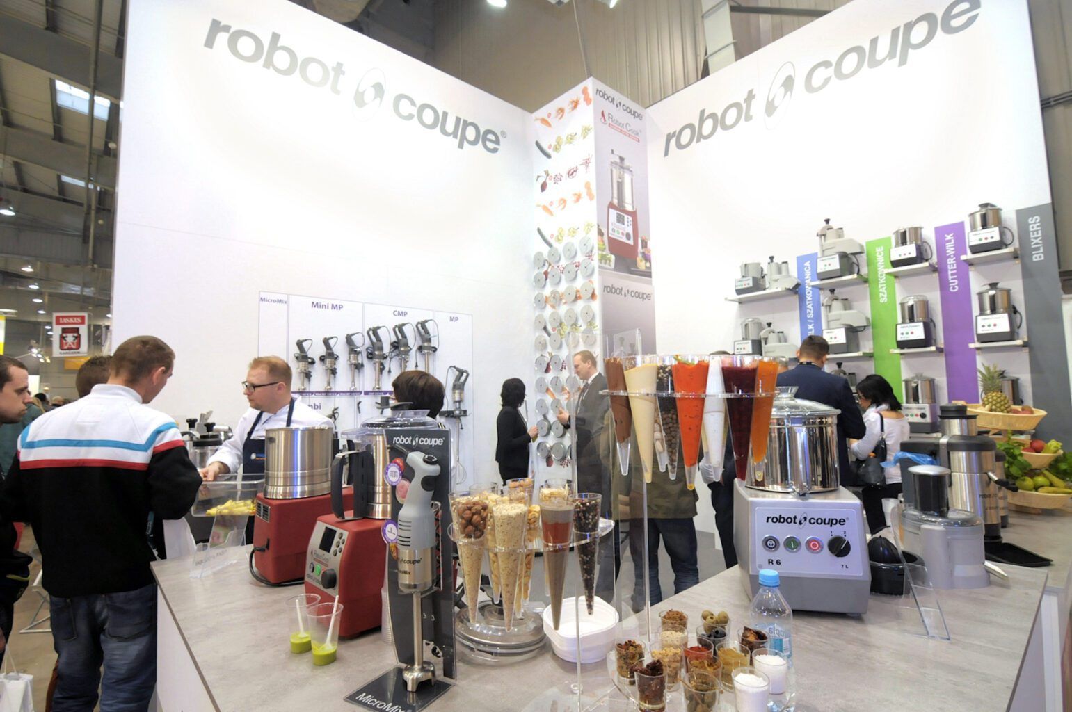 Stoisko targowe prezentuje sprzęt kuchenny Robot Coupe. Odwiedzający wchodzą w interakcję wokół lad z robotami kuchennymi, blenderami i próbkami smoothie. Biała, nowoczesna przestrzeń posiada duże znaki firmowe-projekt i nadzór trksystem.com autorstwa Tatjany Kobuszewskiej. projekt i nadzór Tatjana Kobuszewska trksystem.com