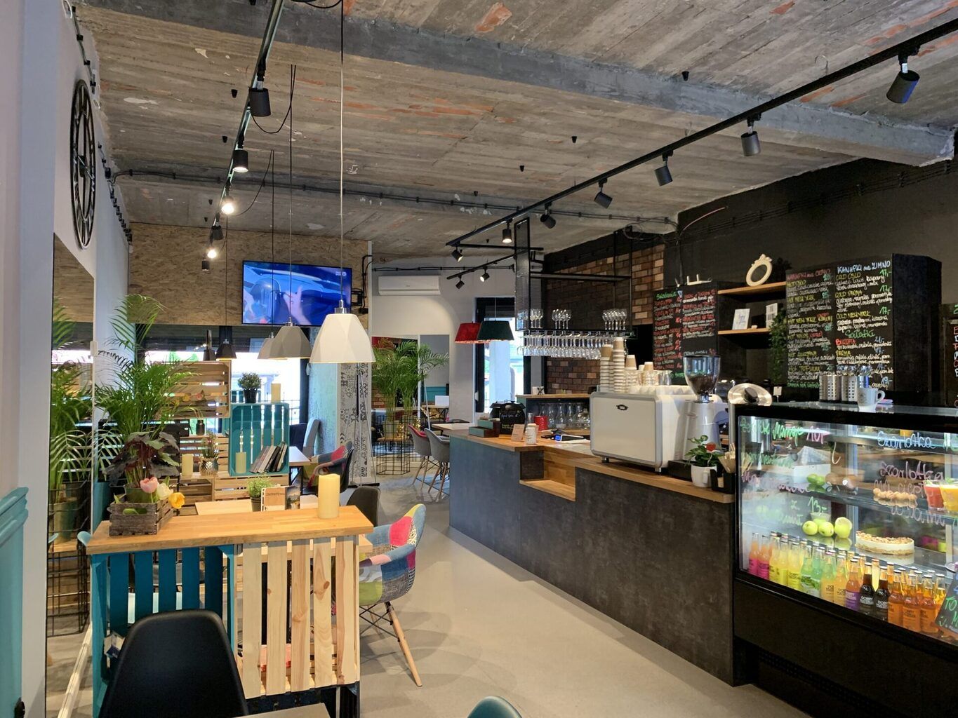 Nowoczesne wnętrze Café Lura z drewnianymi i industrialnymi elementami, kolorowymi krzesłami, roślinami doniczkowymi, kredowym menu na ścianie, ladą z ekspresem do kawy i lodówką z różnymi napojami i deserami. projekt i nadzór Tatjana Kobuszewska trksystem.com