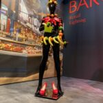 Błyszczący czarny manekin stoi na wystawie w jaskrawoczerwonych szpilkach, ozdobiony kolorowymi akcesoriami ze sztucznych owoców. Za nim znajduje się mural przedstawiający rynek spożywczy i czerwony napis "BÄRO Retail Lighting". Projekt i nadzór trksystem.com by Tatjana Kobuszewska. projekt i nadzór Tatjana Kobuszewska trksystem.com