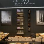 Ekspozycja stoiska Fine Dine na Kongresie Szefów Kuchni FineDine przedstawia szkło, talerze i sztućce na drewnianych stołach. Na czarnej ścianie znajdują się półki z kieliszkami, zdjęcia promocyjne nakryć stołowych oraz logo Fine Dine nad boksami poniżej. projekt i nadzór Tatjana Kobuszewska trksystem.com