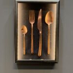 Oprawiony w ramkę ekspozytor prezentuje cztery kawałki sztućców w kolorze miedzi - łyżeczkę, widelec, nóż i łyżkę stołową zamontowane pionowo na ciemnym tle. Ten elegancki element przypomina styl widoczny na wydarzeniach Kongres szefów kuchni FineDine. projekt i nadzór Tatjana Kobuszewska trksystem.com