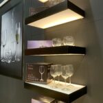 Podświetlane pływające półki na szarej ścianie eksponują puste kieliszki do wina i zestawy szkła Volare. Nieopodal zamontowano duże zdjęcie szkła, prezentowane na Kongresie szefów kuchni FineDine, przedstawiające nalewanie wody do szklanki. projekt i nadzór Tatjana Kobuszewska trksystem.com