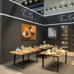 Stoisko targowe z brandingiem Fine Dine na Kongresie szefów kuchni ma czarne ściany, duże zdjęcia jedzenia i szkła oraz stoły z kolorowymi talerzami, miskami, szklankami i sztućcami. Półki z zastawą stołową ustawione są wzdłuż ścian pośród roślin doniczkowych. projekt i nadzór Tatjana Kobuszewska trksystem.com