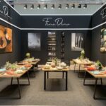 Nowoczesne stoisko wystawiennicze Fine Dine na Kongresie Szefów Kuchni z czterema drewnianymi stołami zastawionymi talerzami, szklankami i sztućcami. Stoisko ma ciemne ściany, elegancki branding, duże zdjęcia restauracji i półki z uporządkowaną zastawą stołową. projekt i nadzór Tatjana Kobuszewska trksystem.com