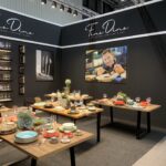Stylowy showroom prezentuje kolorowe naczynia, filiżanki i szklanki na drewnianych stołach. Na czarnych ścianach widnieje napis "Fine Dine", półki z przyborami kuchennymi oraz duże zdjęcie szefa kuchni serwującego potrawy - idealna inspiracja dla każdego szefa kuchni FineDine. W tle widoczne są dwie osoby. projekt i nadzór Tatjana Kobuszewska trksystem.com