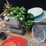 Drewniany stół zastawiony jest kolorowymi talerzami, miskami, szklankami i sztućcami. W centrum rustykalny element centralny z ułożonych w stos betonowych bloków z roślinami i drewnem dryfującym dodaje uroku - idealne ustawienie inspirowane przez Kongres szefów kuchni FineDine. projekt i nadzór Tatjana Kobuszewska trksystem.com