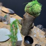 Drewniany stół z talerzami, szklankami i miskami ma centralny element w postaci pnia drzewa pokrytego mchem i paprociami - idealny na imprezę Kongres szefów kuchni FineDine, tworząc rustykalną, inspirowaną naturą aranżację jadalni. projekt i nadzór Tatjana Kobuszewska trksystem.com