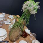 Drewniany element centralny z kaskadą zielonych winorośli i białych omszałych kwiatów zdobi drewniany stół jadalny FineDine z eleganckimi białymi naczyniami, miskami, przyborami kuchennymi, szklankami i filiżankami - idealny na spotkanie Kongres szefów kuchni. projekt i nadzór Tatjana Kobuszewska trksystem.com