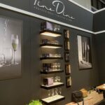 Stoisko targowe prezentuje branding Fine Dine Professional Tabletop Bar, promujący Kongres szefów kuchni FineDine. Półki prezentują szklane naczynia i pudełka z zastawą stołową, a oprawione zdjęcia wiszą nad ladą z talerzami, zieloną rośliną i torbami. projekt i nadzór Tatjana Kobuszewska trksystem.com