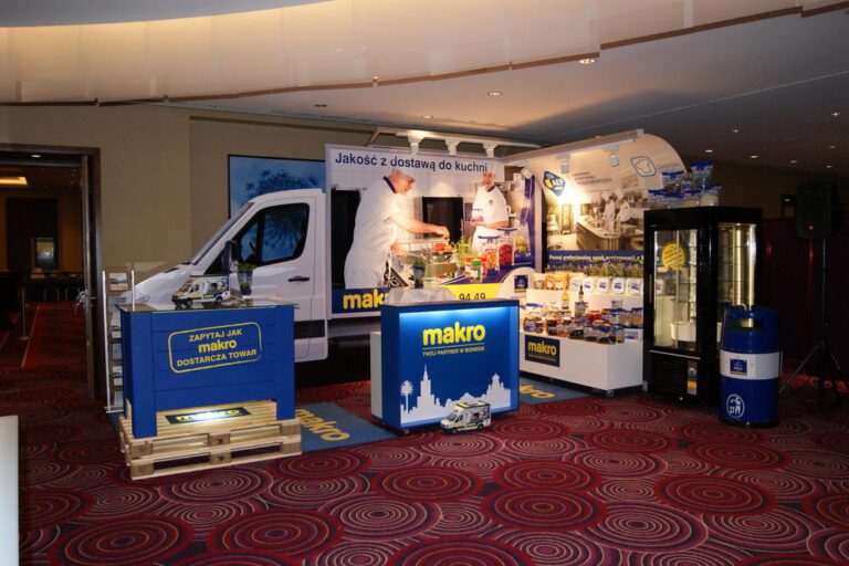 Stoisko targowe z brandingiem „makro”, zaprojektowane przez projekt i nadzór trksystem.com, obejmuje furgonetkę, szefa kuchni przygotowującego jedzenie, eksponowane produkty spożywcze, lodówkę, materiały promocyjne i duże polskie tło ustawione na czerwonym dywanie. projekt i nadzór Tatjana Kobuszewska trksystem.com