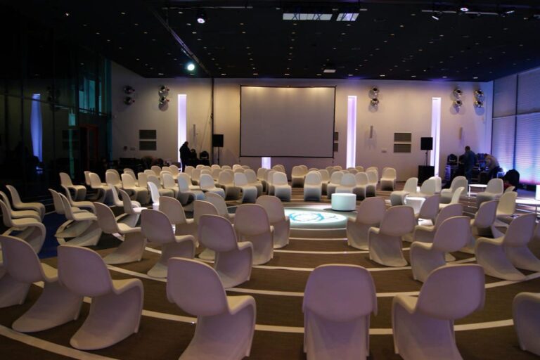 Duża nowoczesna sala konferencyjna z białymi krzesłami ustawionymi w koncentrycznych kręgach zwróconymi w stronę centralnego obszaru i dużym ekranem projekcyjnym, zaprojektowana w ramach projektu i nadzór trksystem.com przez Tatjanę Kobuszewską. Przyćmione reflektory i niebieskie akcenty tworzą nastrój. projekt i nadzór Tatjana Kobuszewska trksystem.com