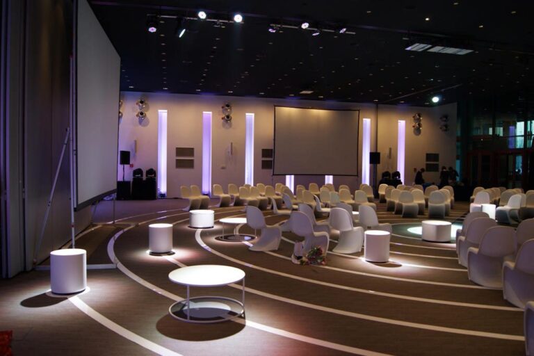 Nowoczesna sala konferencyjna z zakrzywionymi rzędami białych krzeseł i małymi okrągłymi stołami naprzeciwko dwóch dużych ekranów projekcyjnych, oświetlonych fioletowymi i białymi światłami. Zaprojektowana przez Tatjanę Kobuszewską, projekt i nadzór trksystem.com, charakteryzuje się współczesnym wystrojem i wysokimi sufitami. projekt i nadzór Tatjana Kobuszewska trksystem.com