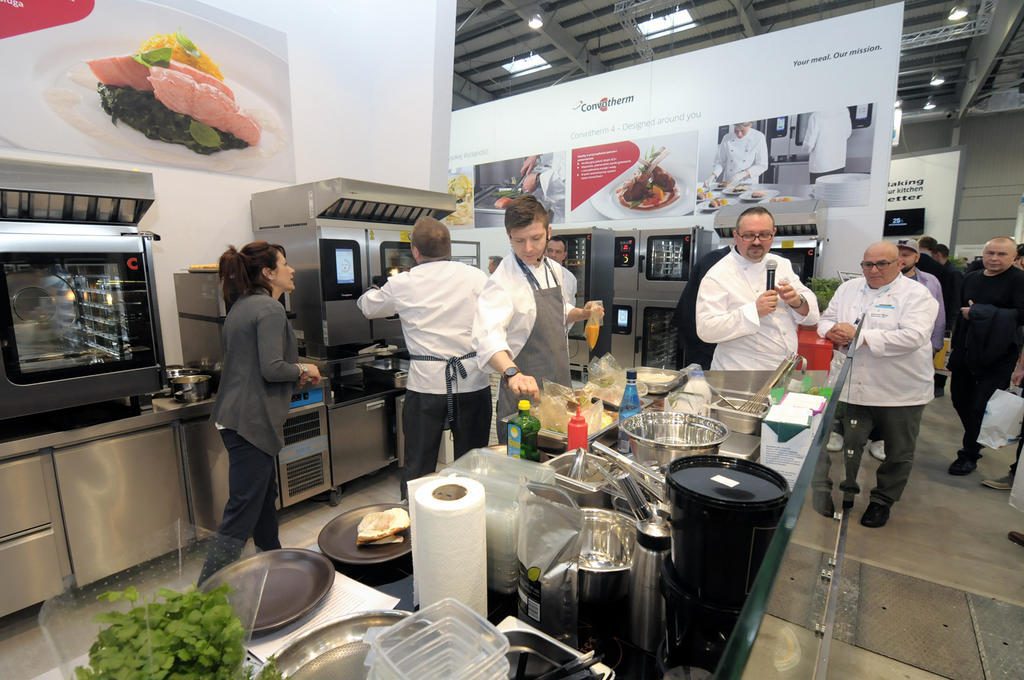 Stoisko Welbilt/ Manitowoc targi EurogastroKucharze w białych uniformach pracują razem w ruchliwej, nowoczesnej kuchni ze sprzętem ze stali nierdzewnej. Dwóch mężczyzn po prawej trzyma talerze, podczas gdy inni przygotowują jedzenie. projekt i nadzór Tatjana Kobuszewska trksystem.com