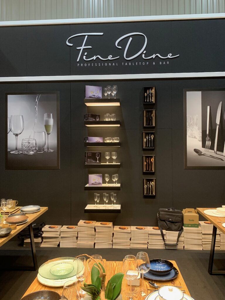 Ekspozycja marki Fine Dine na Kongresie Szefów Kuchni obejmuje szkło, talerze i sztućce wyeksponowane na drewnianych stołach, pod którymi ułożono stosy katalogów. Na czarnej ścianie widnieje logo Fine Dine, regały, zestawy sztućców oraz dwa duże zdjęcia nakryć stołowych. projekt i nadzór Tatjana Kobuszewska