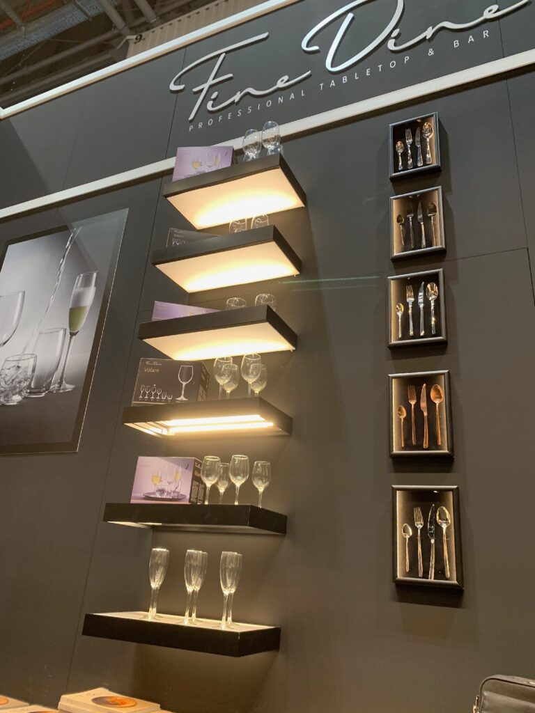 Na ścianie ekspozycyjnej znajdują się podświetlane półki z różnymi kieliszkami do wina, a nad nimi widnieje napis „Fine Dine Professional Tabletop Bar”. Po prawej stronie, w pionowych rzędach, starannie ułożone są zestawy sztućców w pudełkach z serii „Kongres szefów kuchni FineDine”. projekt i nadzór Tatjana Kobuszewska stoisko na kongresie szefów kuchni