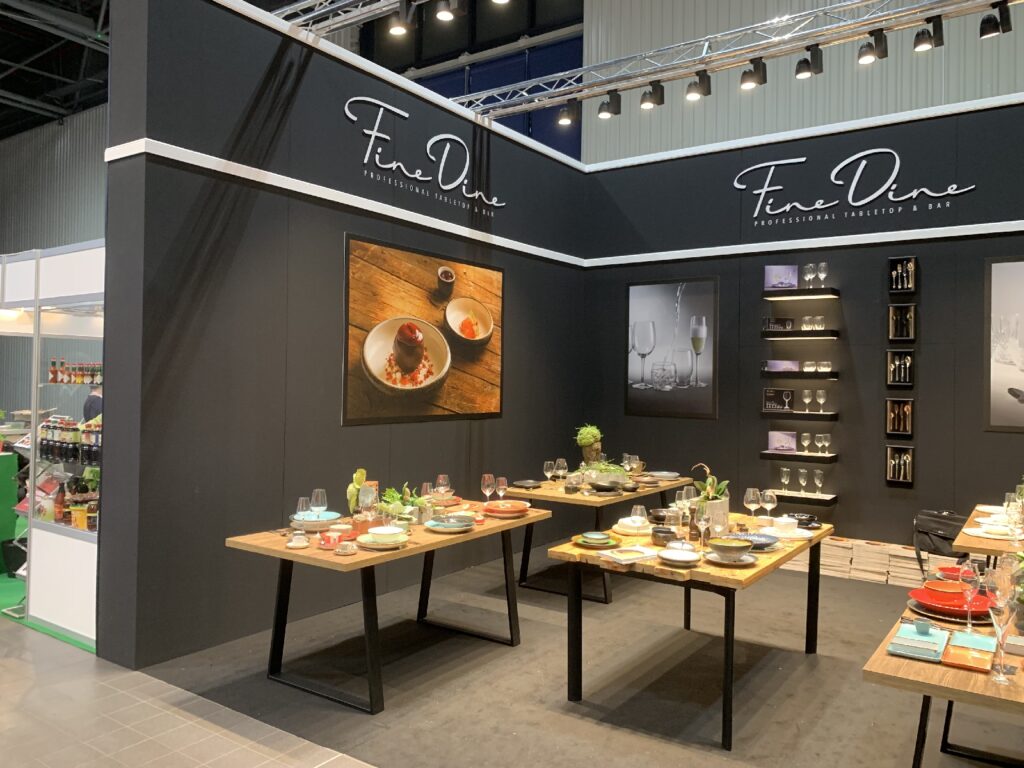 Stoisko targowe firmy „Fine Dine” na Kongresie Szefów Kuchni charakteryzuje się czarnymi ścianami, elegancką zastawą stołową, ekspozycjami szkła oraz nakryciami na drewnianych stołach. Duże zdjęcia potraw i reflektory punktowe tworzą stylową, nowoczesną atmosferę. projekt i nadzór Tatjana Kobuszewska